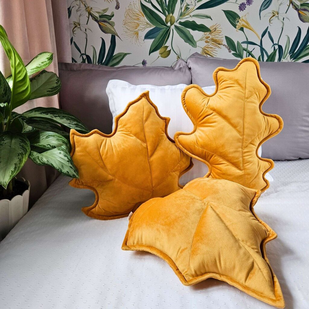Zestaw trzech musztardowych poduszek dekoracyjnych w kształcie liści (klonu, dębu i brzozy) ułożonych na jasnej kanapie. Welurowe dodatki tworzą przytulny klimat w słonecznym salonie i są gotowym pomysłem na ocieplenie wnętrza bez remontu.