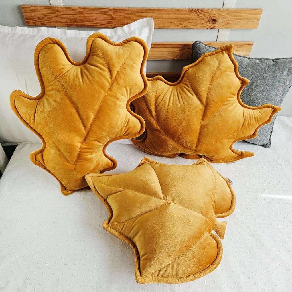 Zestaw trzech musztardowych poduszek dekoracyjnych w kształcie liści (klonu, dębu i brzozy) ułożonych na jasnej kanapie. Welurowe dodatki tworzą przytulny klimat w słonecznym salonie i są gotowym pomysłem na ocieplenie wnętrza bez remontu.