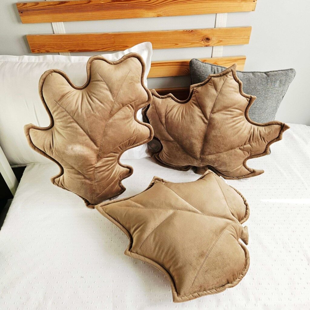 Zestaw trzech poduszek dekoracyjnych w kształcie liści (klonu, dębu i brzozy) ułożonych na szarej kanapie. Welurowe dodatki tworzą przytulny klimat w słonecznym salonie i są gotowym pomysłem na ocieplenie wnętrza bez remontu.