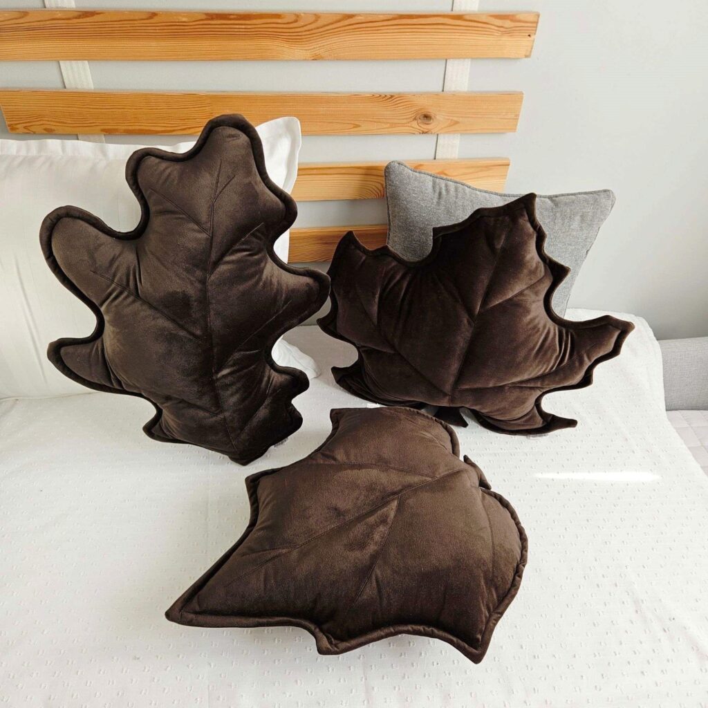 Zestaw trzech poduszek dekoracyjnych w kształcie liści (klonu, dębu i brzozy) ułożonych na szarej kanapie. Welurowe dodatki tworzą przytulny klimat w słonecznym salonie i są gotowym pomysłem na ocieplenie wnętrza bez remontu.