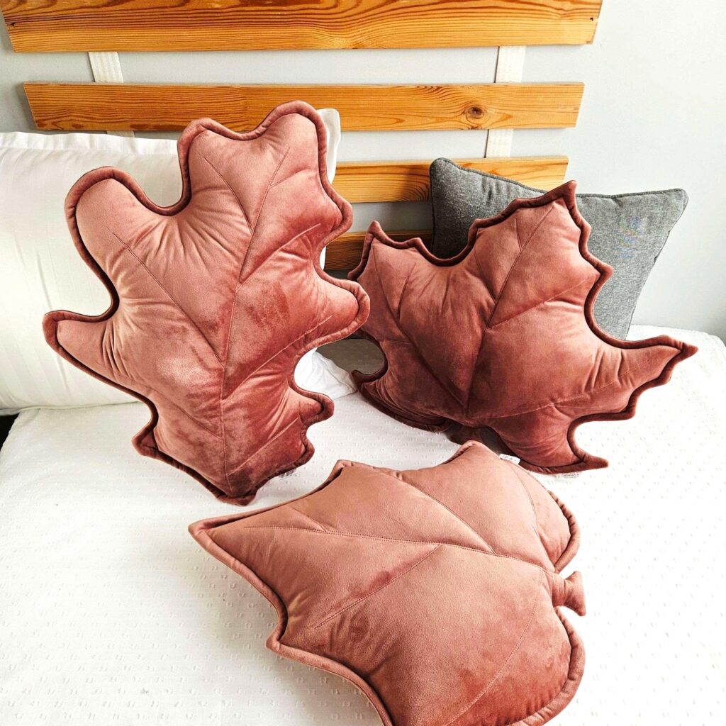 Zestaw trzech poduszek dekoracyjnych w kształcie liści (klonu, dębu i brzozy) ułożonych na szarej kanapie. Welurowe dodatki tworzą przytulny klimat w słonecznym salonie i są gotowym pomysłem na ocieplenie wnętrza bez remontu.