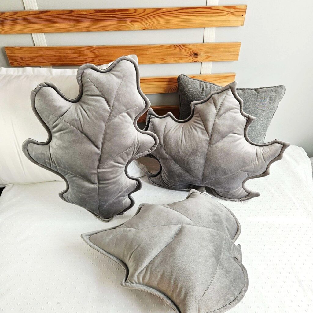 Zestaw trzech poduszek dekoracyjnych w kształcie liści (klonu, dębu i brzozy) ułożonych na szarej kanapie. Welurowe dodatki tworzą przytulny klimat w słonecznym salonie i są gotowym pomysłem na ocieplenie wnętrza bez remontu.