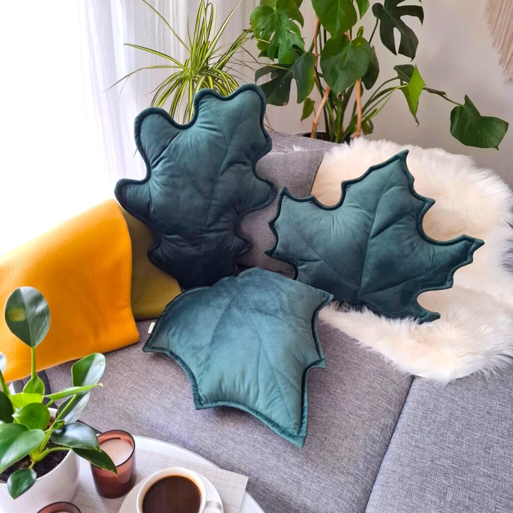 Zestaw trzech poduszek dekoracyjnych w kształcie liści (klonu, dębu i brzozy) ułożonych na szarej kanapie. Welurowe dodatki tworzą przytulny klimat w słonecznym salonie i są gotowym pomysłem na ocieplenie wnętrza bez remontu.
